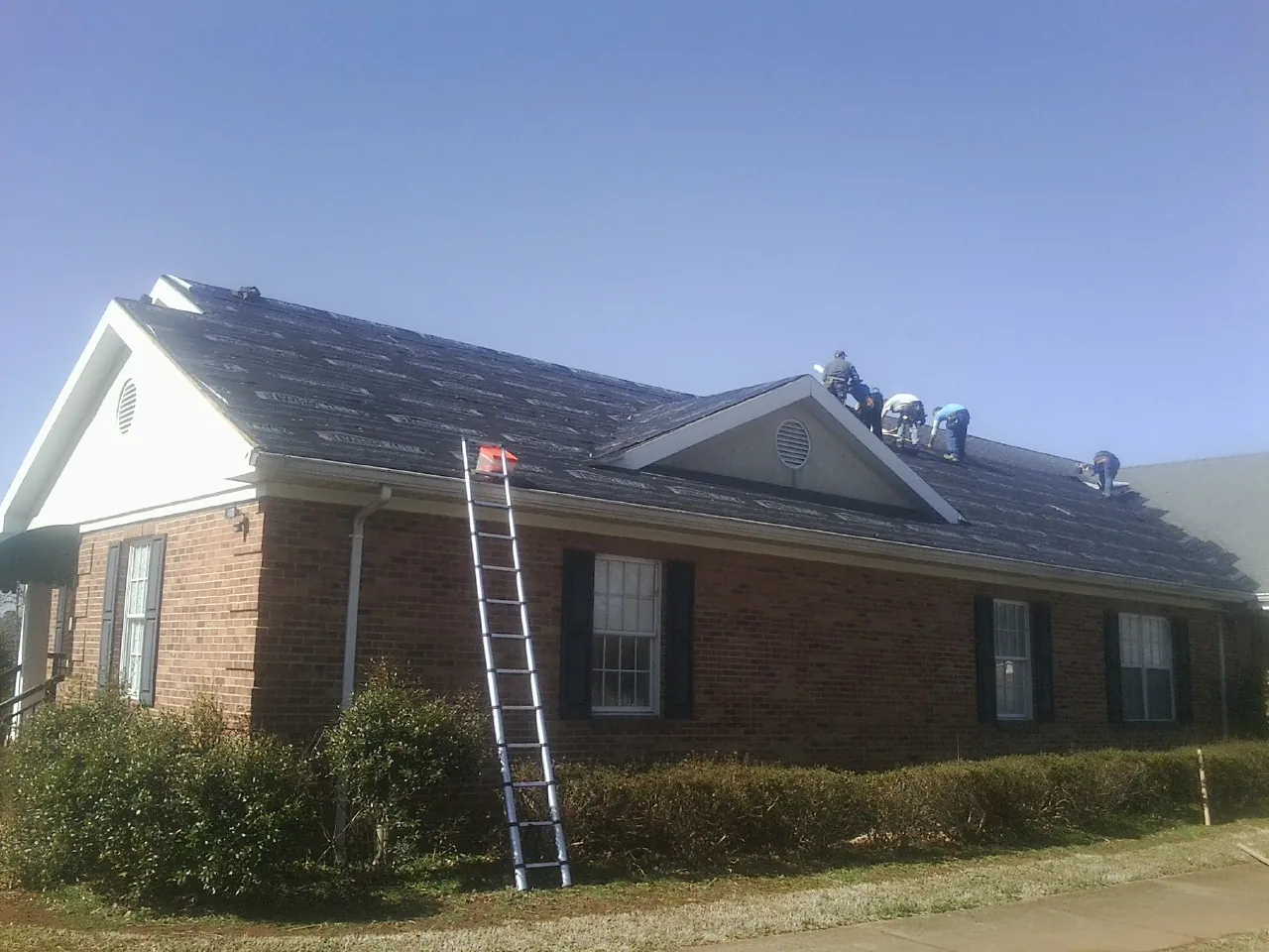 Crew tearing shingles off a brick ranch roof with a ladder set — Metal Roof Repair work by our crew in Remington